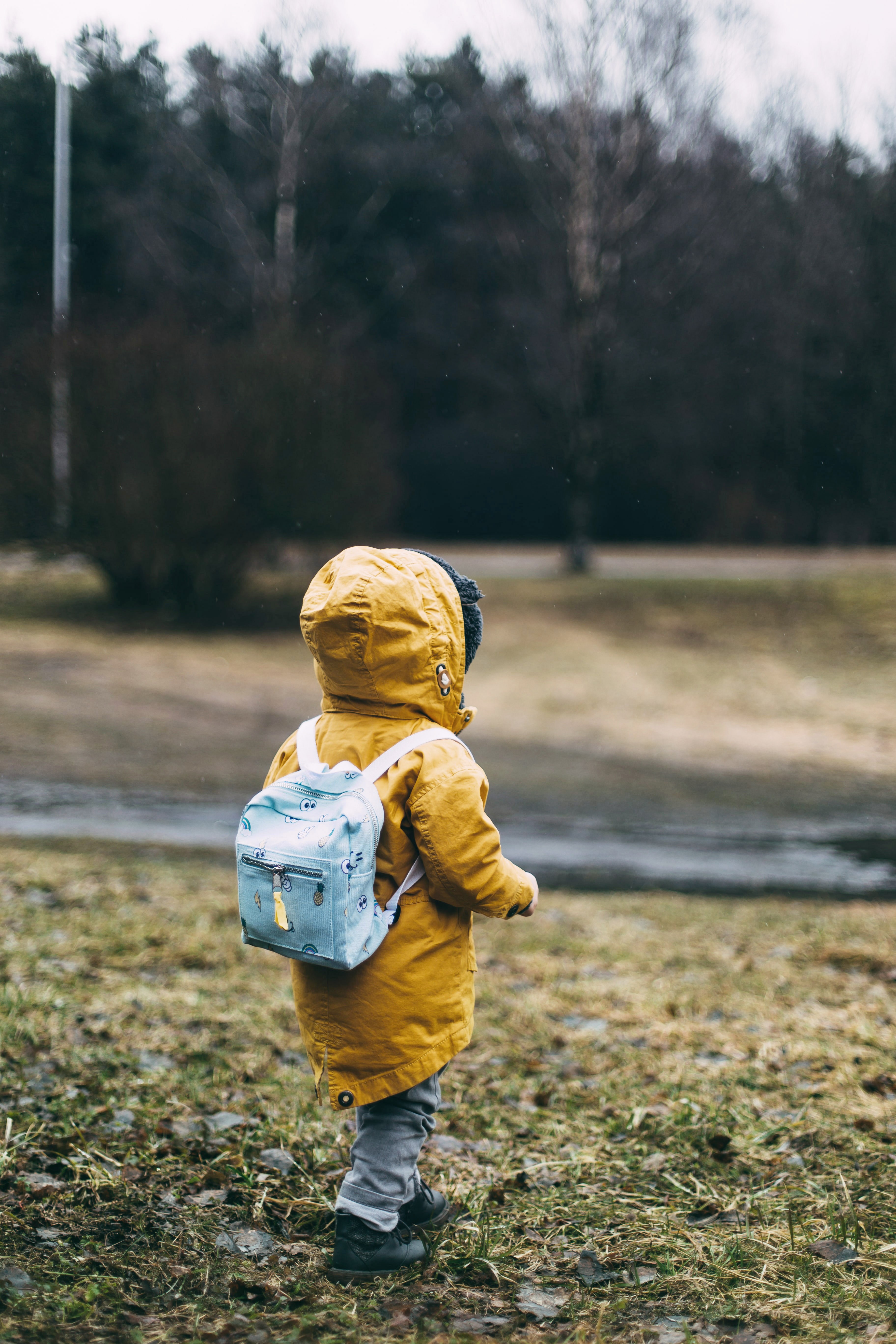 Ett barn som går ensam på en äng. Barnet har en gul jacka på sig och en ljusblå ryggsäck. I bakgrunden syns ett skogsparti. Vädret är grått.  