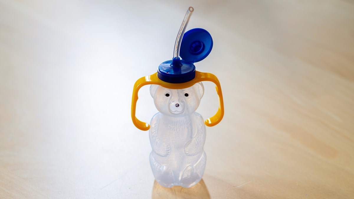 Flaska i genomskinlig plast i form av en björn med gula handtag och ett blått lock med integrerad sugrör. 